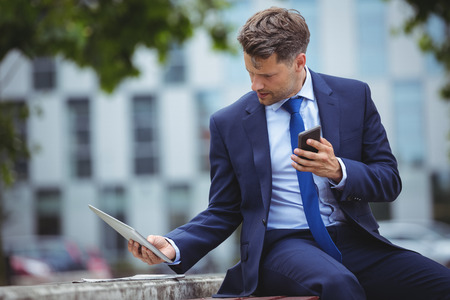 Handsome businessman holding mobile phone while using digital tablet outside officeの写真素材