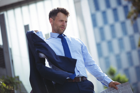Thoughtful businessman holding blazer and newspaper near office buildingの写真素材