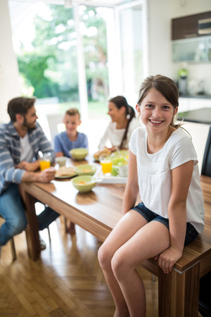 Portrait of girl sitting on dining table while family having breakfast in background at homeの写真素材