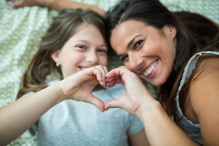 Mother and daughter making heart shape from hand while lying on bed at homeの写真素材