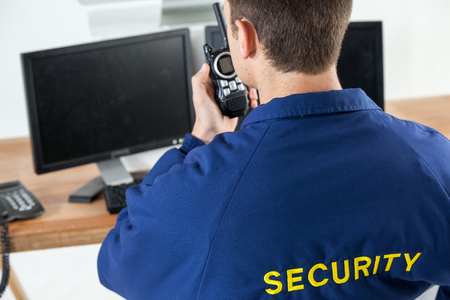 Rear view of security officer talking on walkie-talkie while looking at computer monitorsの写真素材