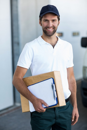 Portrait of happy delivery man holding parcel and clipboard outside the warehouseの写真素材