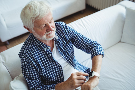 Senior man adjusting a time on smartwatch in living room at homeの写真素材