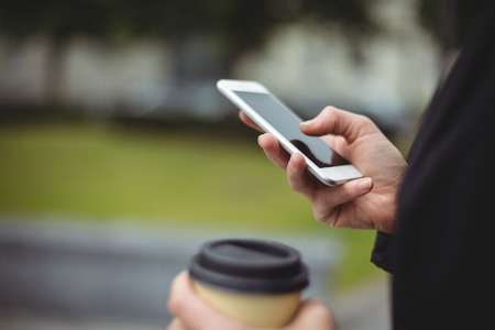 Mid section of businesswoman using mobile phone while holding disposable coffee cup in office campusの写真素材