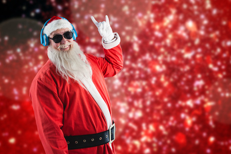 Santa claus showing hand yo sign while listening to music on headphones against white snow and stars on redの写真素材