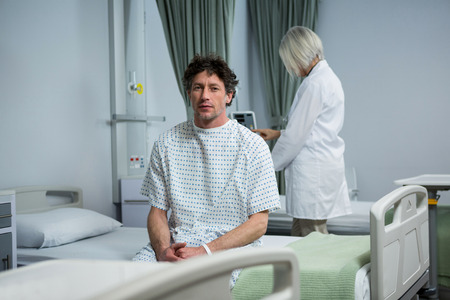 Portrait of patient sitting on bed in ward while doctor standing in backgroundの写真素材