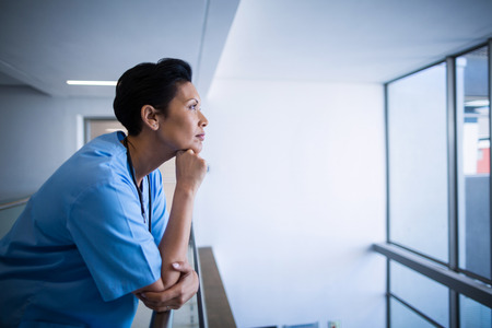 Thoughtful female nurse leaning on railing in corridor of hospitalの写真素材