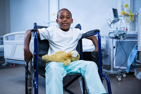 Portrait of boy sitting on wheelchair with teddy bear in ward at hospitalの写真素材