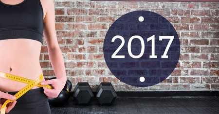 Mid section of woman measuring her waist against 2017 with dumbbells in background