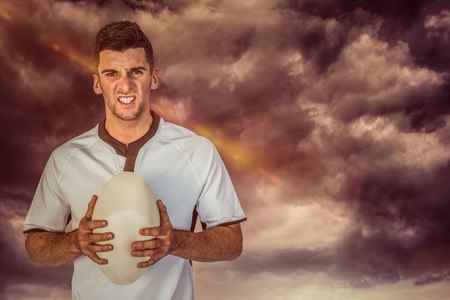3D Portrait of an angry rugby player holding ball against gloomy skyの写真素材