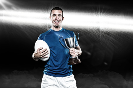 Portrait of smiling rugby player holding trophy and ball against spotlight 3Dの写真素材