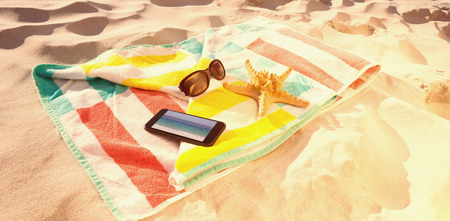 Starfish with sunglasses and mobile phone kept on beach blanket against waters edge at the beachの写真素材