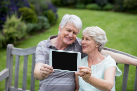 Senior couple sitting on bench and holding digital tablet in lawnの写真素材