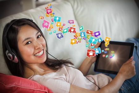 Girl using her tablet pc on the sofa and listening to music smiling at camera against colourful computer applications 3dの写真素材