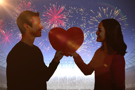 Smiling couple holding red heart shape  against fireworks exploding over football stadiumの写真素材