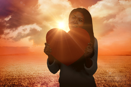 Smiling Asian woman holding paper heart against sunrise over fieldの写真素材