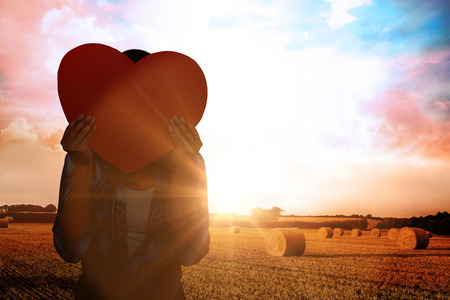 Woman hiding her face behind paper heart against countryside sceneの写真素材