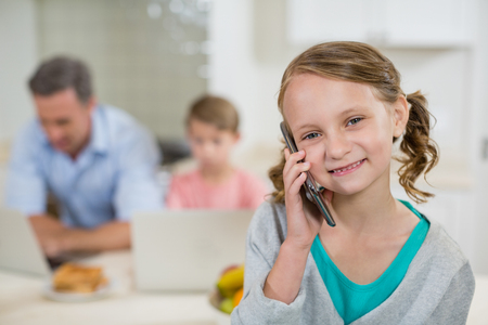 Portrait of cute girl talking on mobile phone in kitchen at homeの写真素材