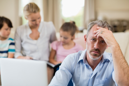 Portrait of tense man with hand on forehead sitting in living room at homeの写真素材