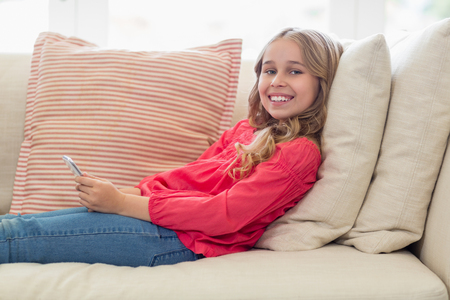 Portrait of smiling girl with mobile phone lying on sofa in living roomの写真素材