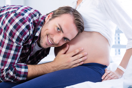 Smiling man listening to pregnant womans belly in ward of hospitalの写真素材