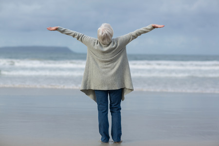 Rear view of senior woman standing with arms outstretched on the beachの写真素材