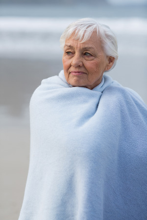Thoughtful senior woman wrapped in shawl on the beachの写真素材