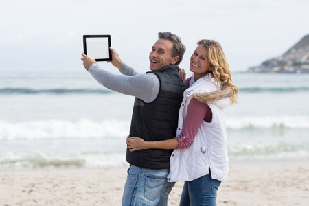 Portrait of mature couple taking selfie with digital tablet on the beachの写真素材