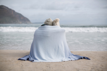 Rear view of mature couple wrapped in blanket on the beachの写真素材
