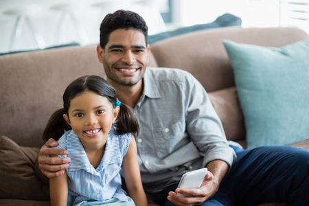 Portrait of father and daughter sitting on sofa in living room at homeの写真素材