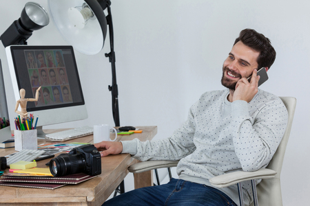 Happy photographer sitting at desk and talking on smartphone in officeの写真素材