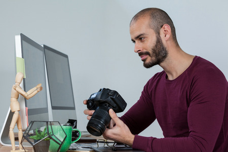 Photographer looking at computer screen while holding camera at desk in officeの写真素材