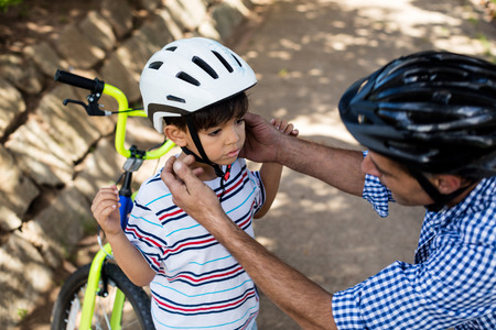 Father assisting son in wearing bicycle helmet in park on a sunny dayの写真素材
