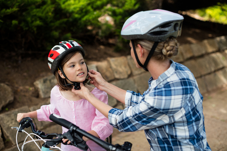 Mother assisting daughter in wearing bicycle helmet in park on a sunny dayの写真素材