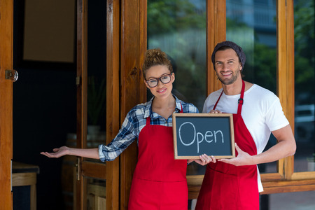 Portrait of smiling waitress and waiter standing with open sign board outside cafÃ©
