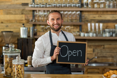 Portrait of smiling waiter standing with open sign board in cafe