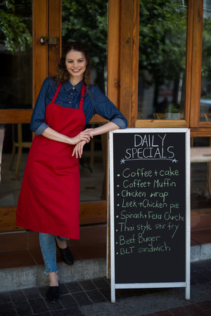 Portrait of smiling waitress leaning on menu board outside the cafeの写真素材