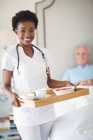 Portrait of smiling nurse with breakfast in tray while senior man lying on bed at homeの写真素材