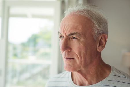 Thoughtful senior man standing next to window at homeの写真素材