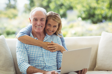 Portrait of smiling senior woman embracing a man in living room while using laptop at homeの写真素材
