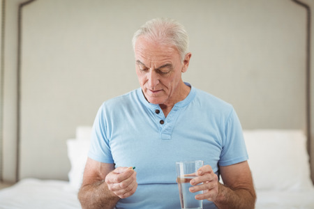 Senior man sitting in bedroom holding medicine and glass of waterの写真素材