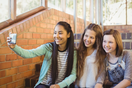 Smiling schoolgirls sitting on the staircase taking selfie with mobile phone at schoolの写真素材