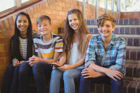 Smiling school students sitting on the staircase using mobile phone at schoolの写真素材