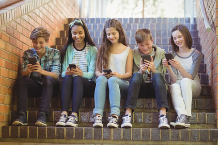 Group of smiling school friends sitting on staircase using mobile phone at schoolの写真素材