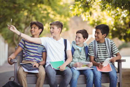 Happy students sitting on bench and taking selfie on mobile phone in school campusの写真素材