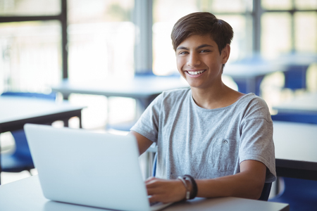 Portrait of happy schoolboy using laptop in classroom at schoolの写真素材