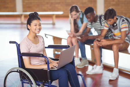 Portrait of disabled schoolgirl using laptop with classmates in background at schoolの写真素材