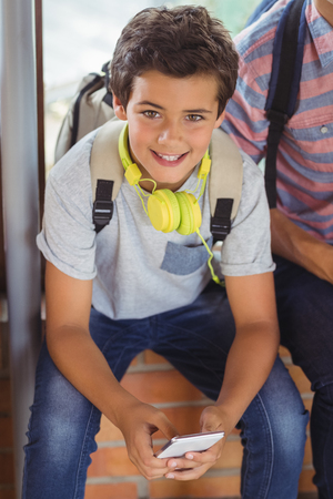Portrait of happy schoolboy sitting on window sill and using mobile phone in corridor at schoolの写真素材