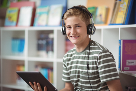 Portrait of happy schoolboy listening music while using digital tablet in libraryの写真素材