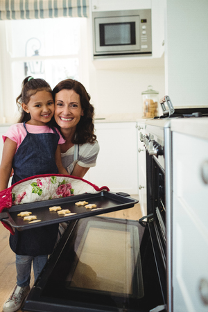 Portrait of mother and daughter holding tray of baked cookies in kitchen at homeの写真素材